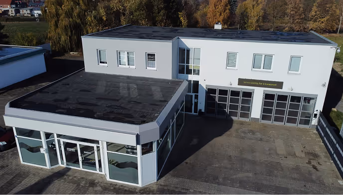 Blick von oben auf ein modernes, zweistöckiges Bürogebäude mit vielen Fenstern und einer großen gepflasterten Fläche davor, umgeben von Bäumen im Herbst.