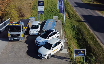 Drei weiße Einsatzfahrzeuge und ein Lastwagen auf einem Parkplatz neben einer Straße und einem Schild mit dem Logo einer Sicherheitsfirma.