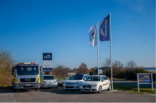 Drei geparkte Autos und ein LKW auf einem Parkplatz vor Fahnen und einem Schild mit Firmenlogo unter blauem Himmel.