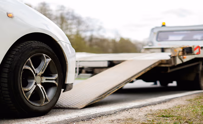 Ein weißes Auto mit einem platten Vorderreifen wird von einem Abschleppwagen mit ausgefahrener Rampe abgeschleppt.