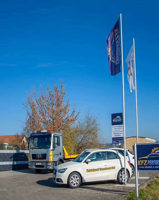 Ein weißes Kombi-Auto mit der Aufschrift 'Werkstattpause? Wir spritzen für Putz!' geparkt vor einem KFZ-Performance-Service mit zwei Fahnen und einem Lastwagen unter klarem blauem Himmel.