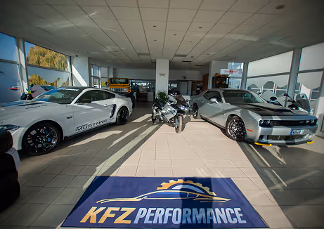 Innenansicht eines Auto-Showrooms mit einem weißen Ford Mustang, einem schwarzen Motorrad und einem grauen Dodge Challenger auf einem Fliesenboden mit einem Teppich, auf dem KFZ Performance steht.