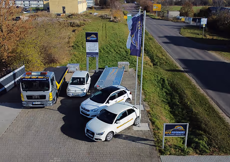 Drei weiße Fahrzeuge auf gepflastertem Parkplatz neben einer Straße mit zwei Fahnen und mehreren Schildern eines Auto- oder Abschleppdienstes.