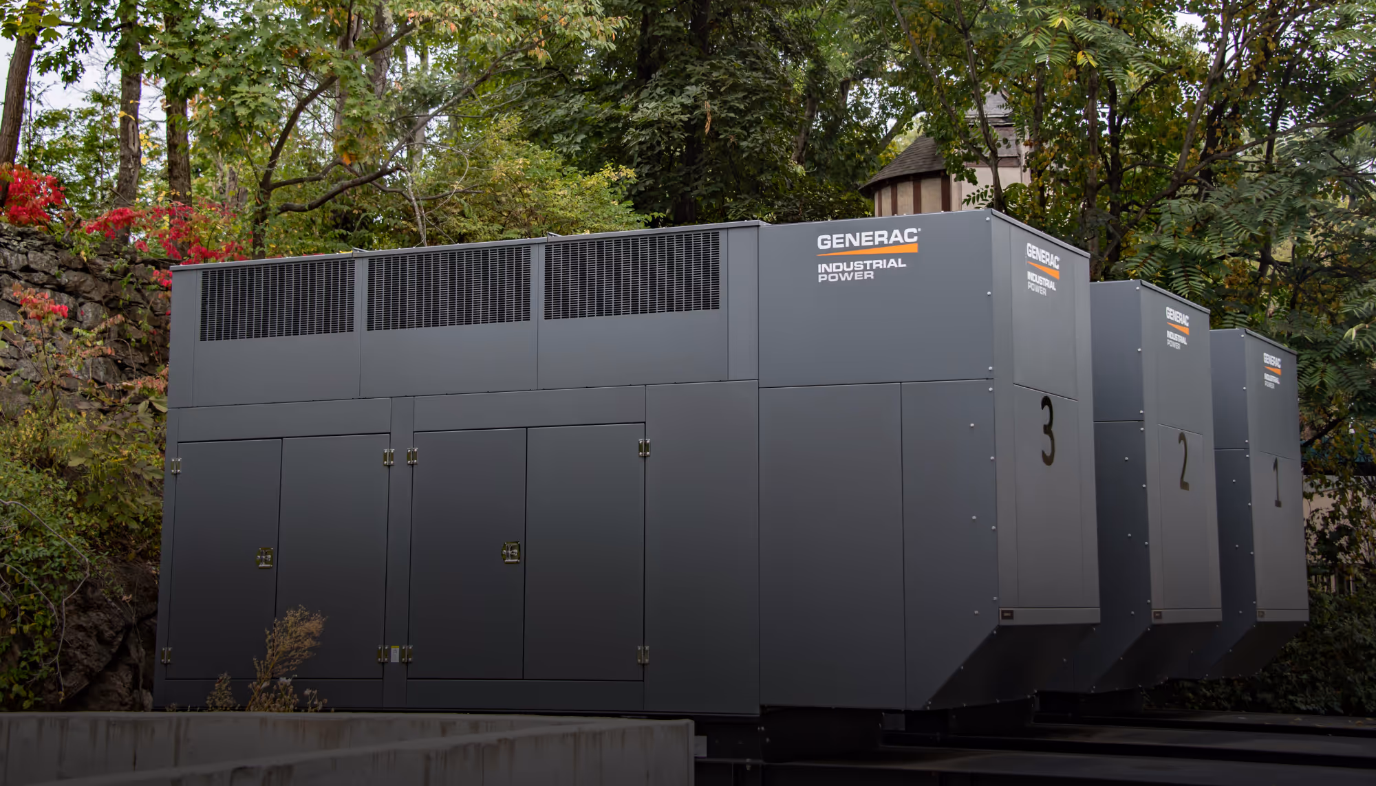 Three large gray industrial power generator units labeled Generac Industrial Power, numbered 3, 2, and 1, placed outdoors near trees and vegetation.