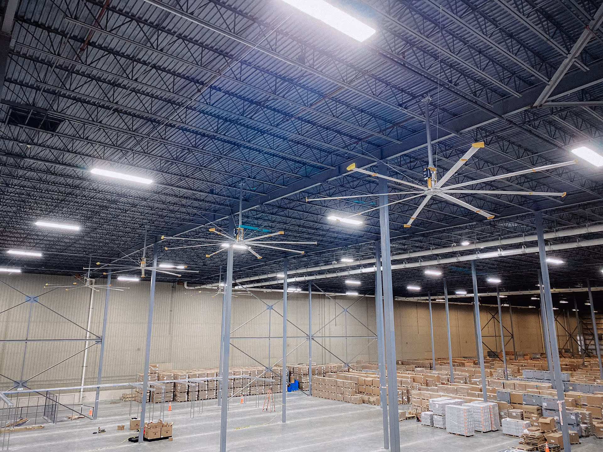 Construction worker wearing a hard hat and safety vest standing on a red ladder looking at an exposed ceiling with ductwork and a ceiling fan.