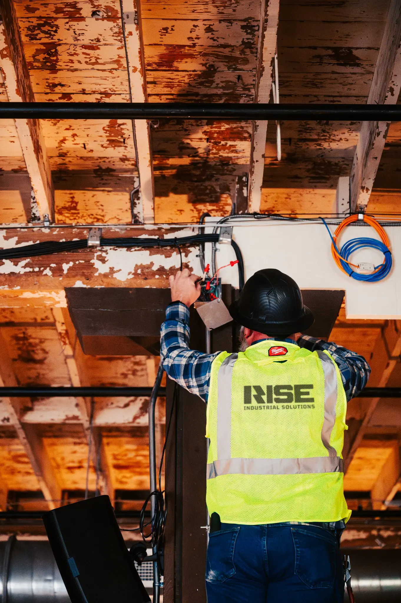 Construction worker in a black hard hat and yellow safety vest using pliers to work on electrical wiring on a wooden ceiling.