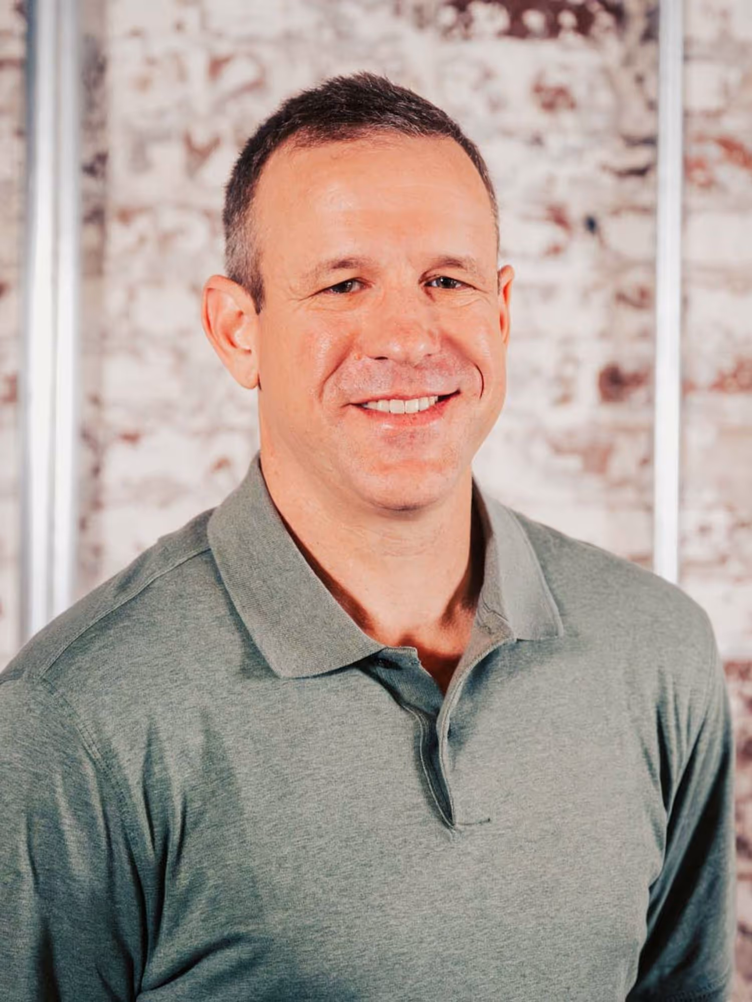 Smiling man wearing a green polo shirt standing in front of a brick wall with metal pipes.