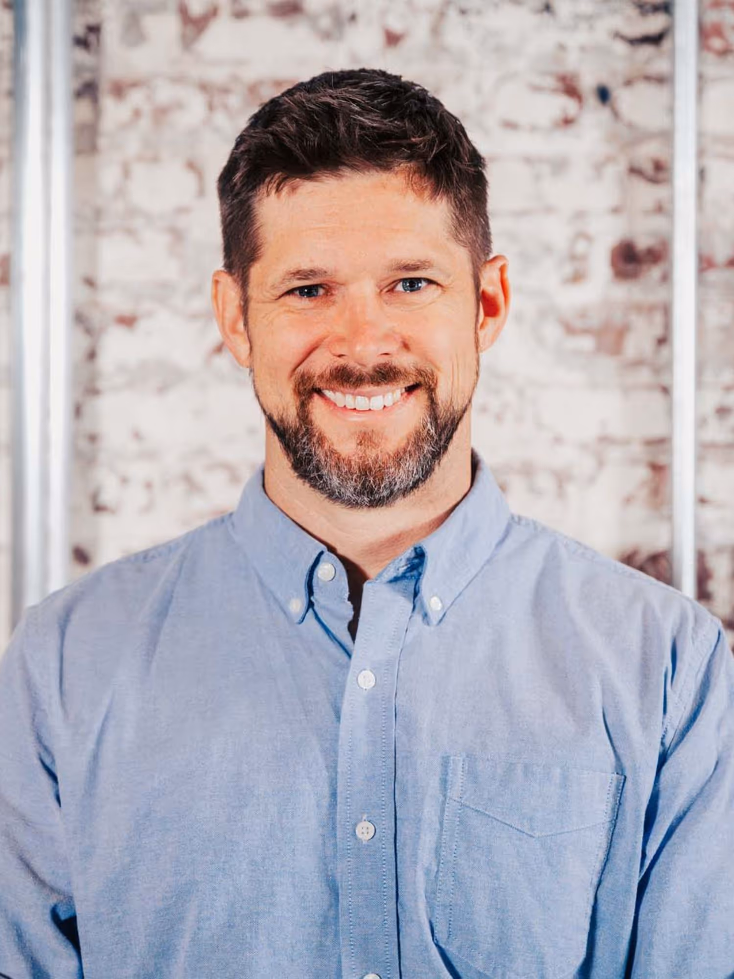 Smiling man with short dark hair and beard wearing a light blue button-up shirt standing in front of a brick wall.