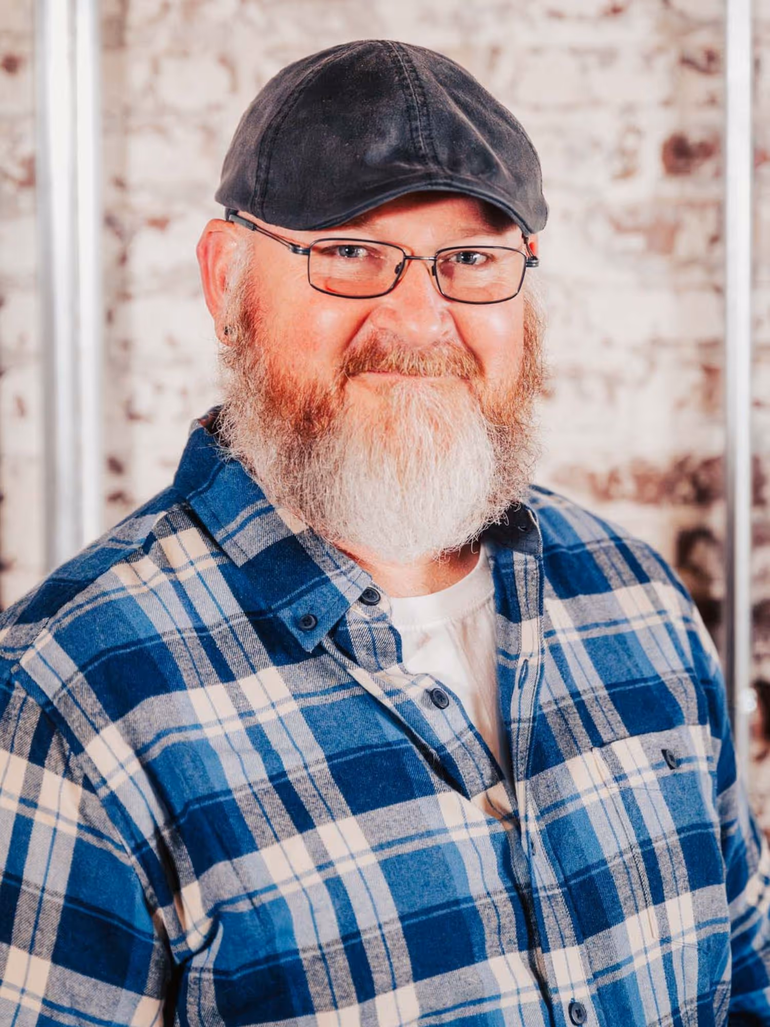 Smiling middle-aged man with a gray and white beard, wearing glasses, a black cap, and a blue plaid shirt, standing in front of a textured brick wall.