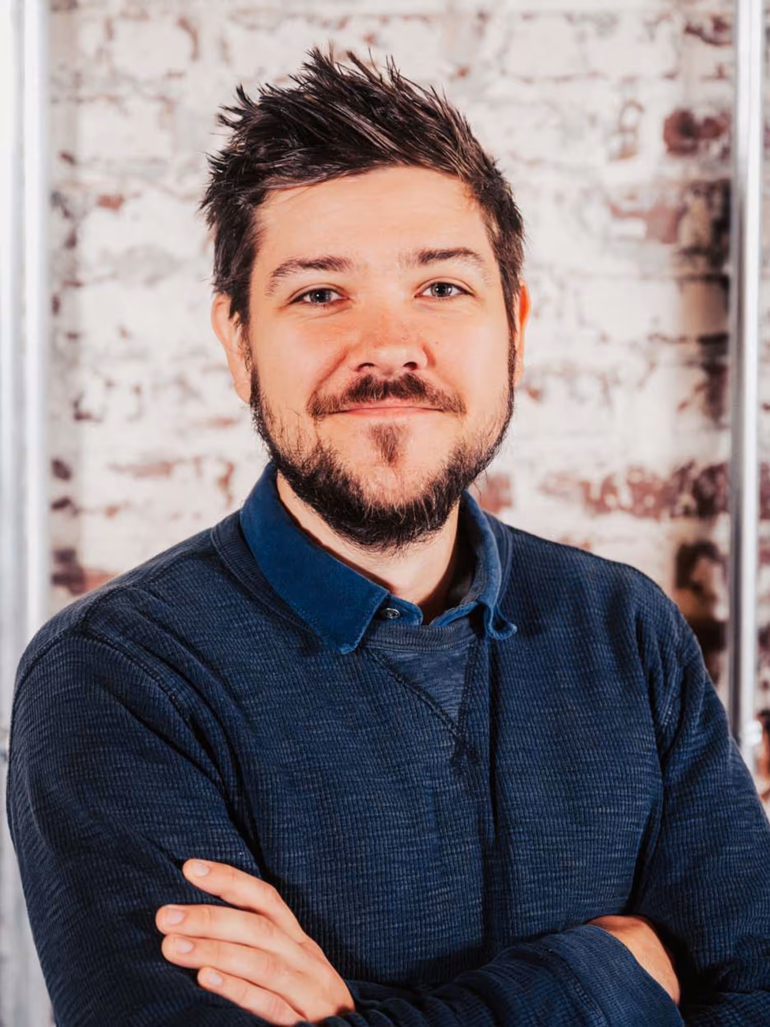 Smiling man with beard and spiked hair wearing a dark blue sweater standing with arms crossed in front of a brick wall.