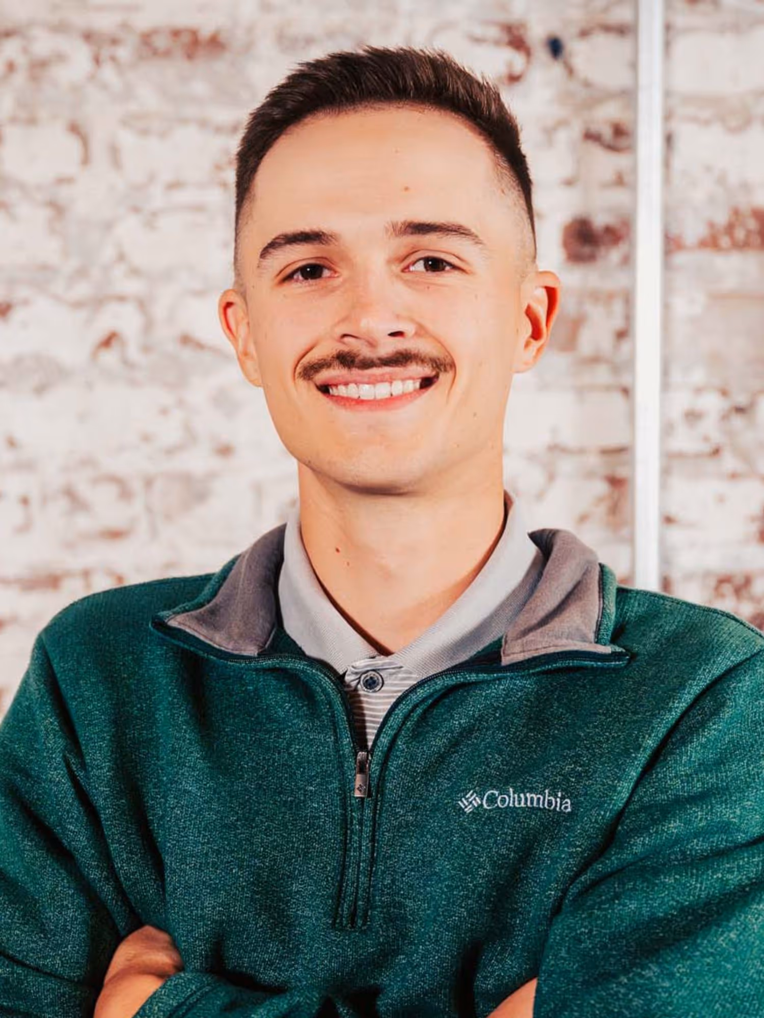 Smiling young man with short hair and mustache wearing a green Columbia pullover sweater standing in front of a brick wall.