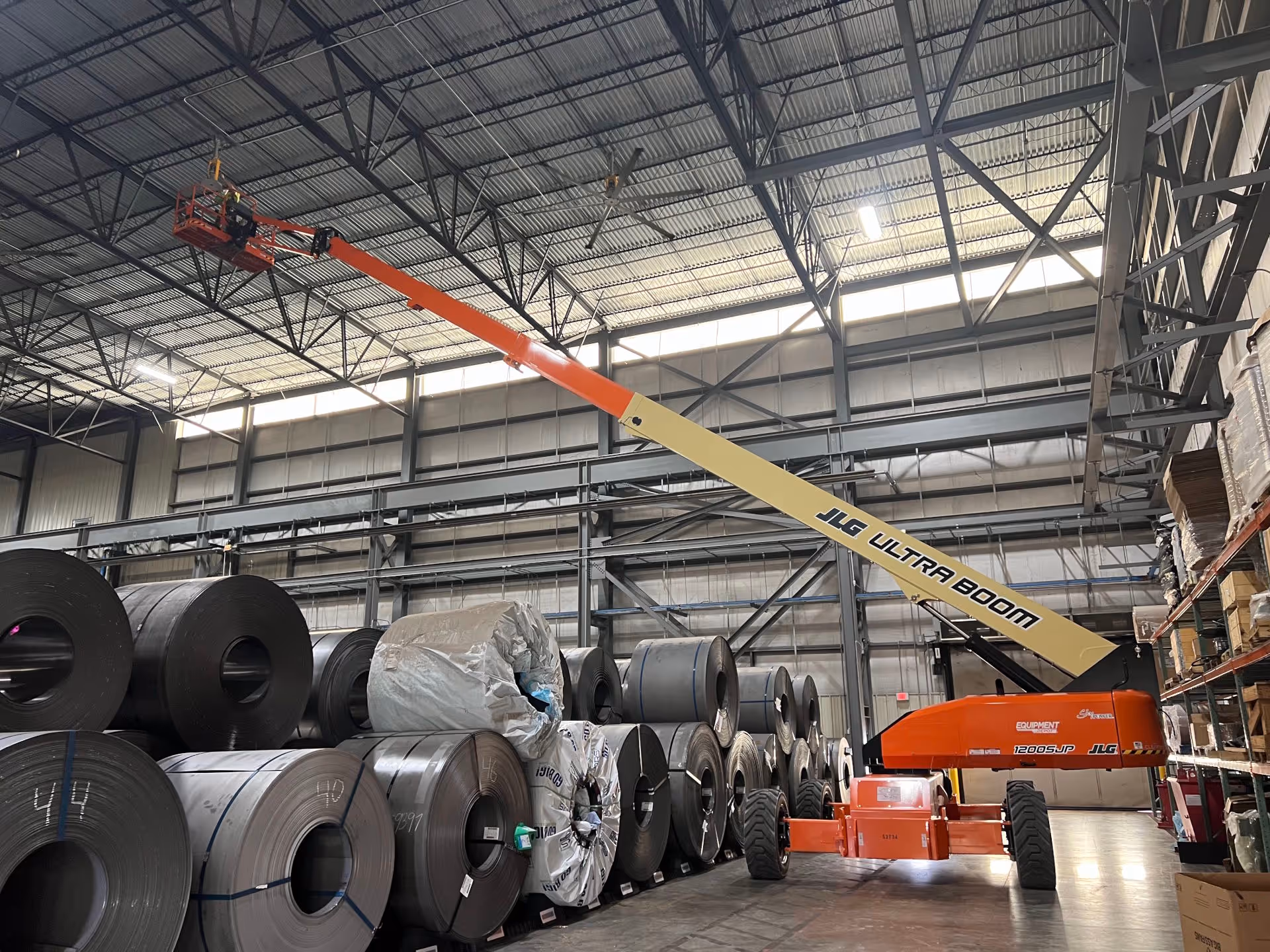 Worker wearing yellow safety vest and white helmet installing or repairing a large ceiling fan in an industrial space with exposed wooden beams.