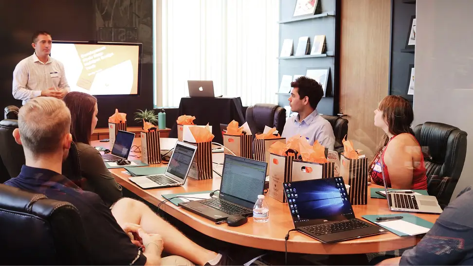 Group of coworkers seated around a conference table listening to a man presenting with a screen in a modern office.