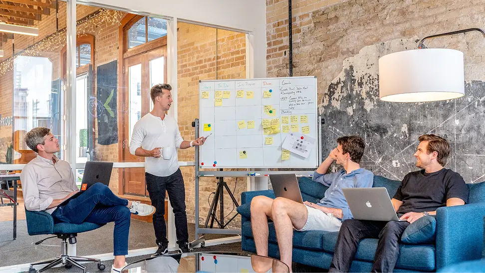 Four young men in a modern office, three seated with laptops and one standing, presenting ideas using a whiteboard with sticky notes.