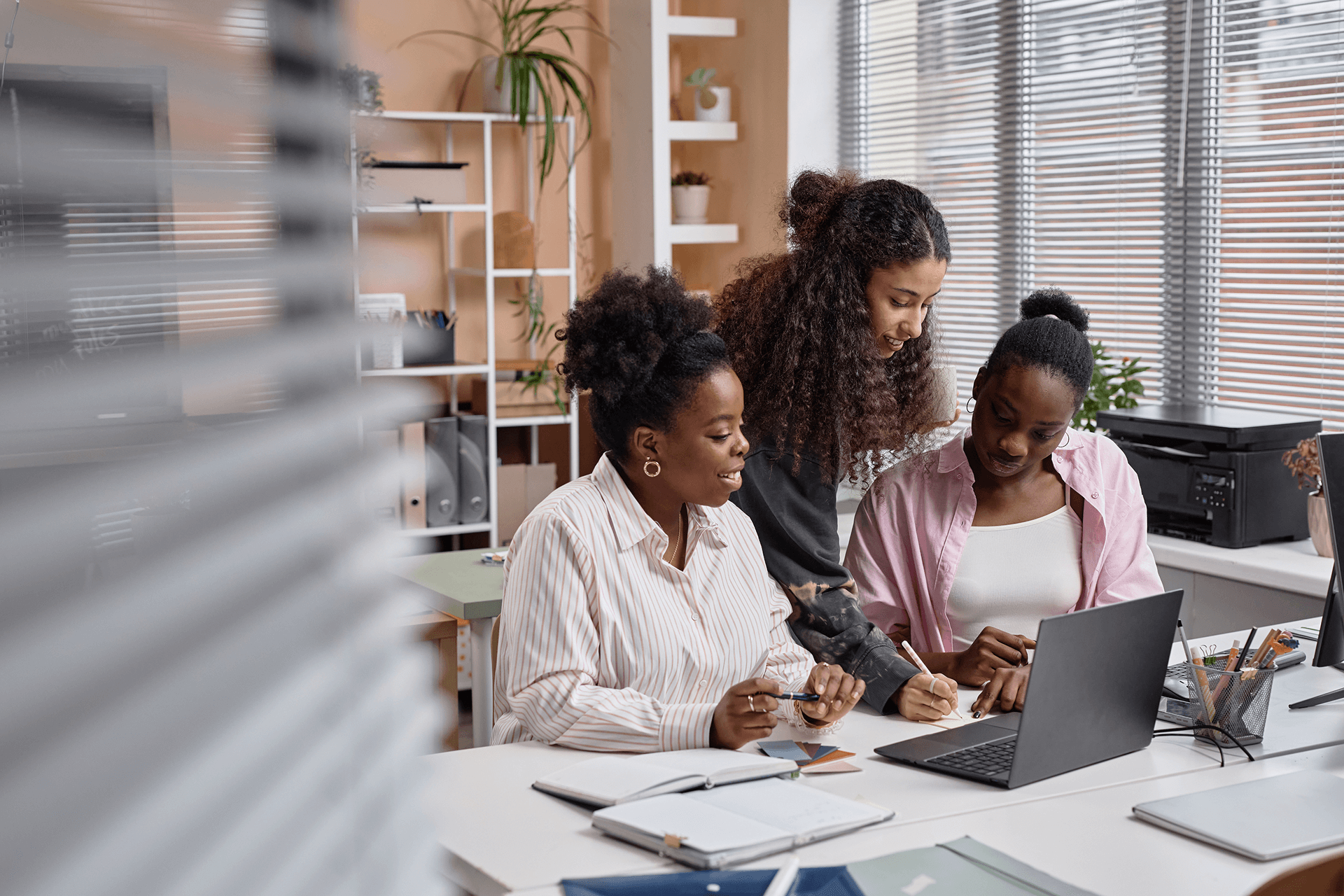 Three women working collaboratively at a desk with a laptop and notebooks in a bright office space.