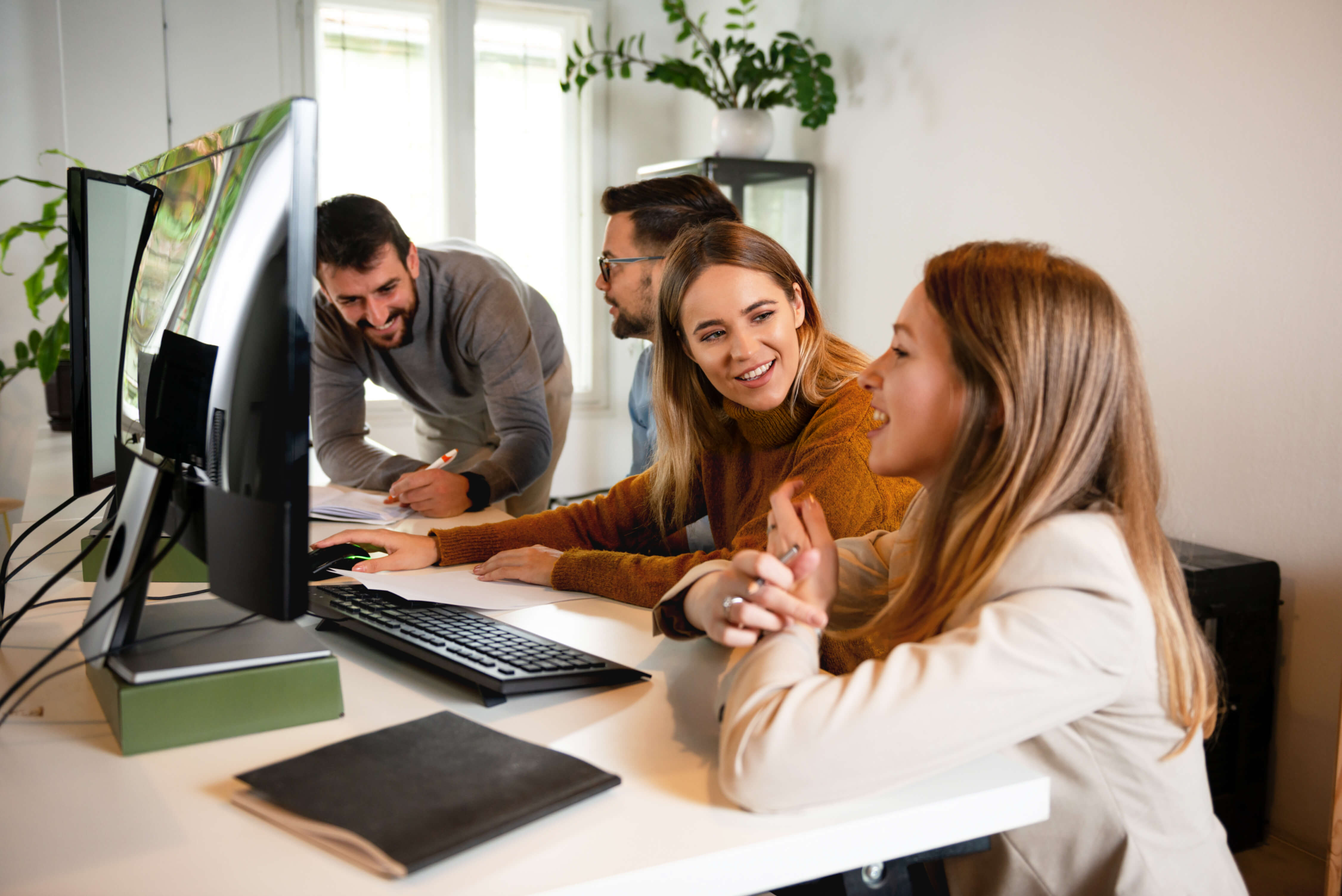 Four coworkers collaborating and smiling around a computer in a bright office.