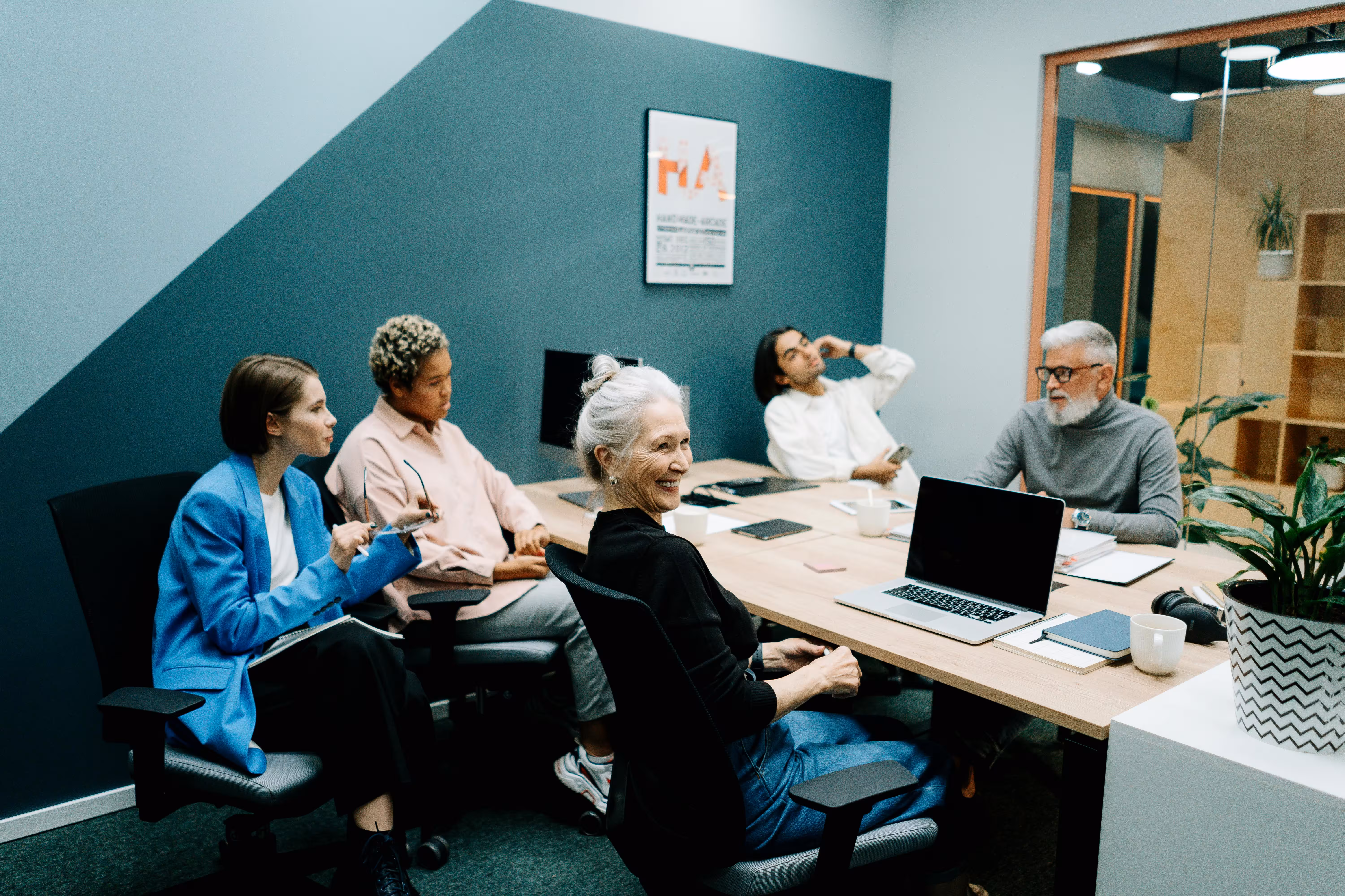 Five diverse professionals having a meeting around a table in a modern office, with laptops, notebooks, and coffee cups.