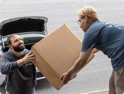 Two men exchanging a large cardboard box near an open car trunk on a paved area.