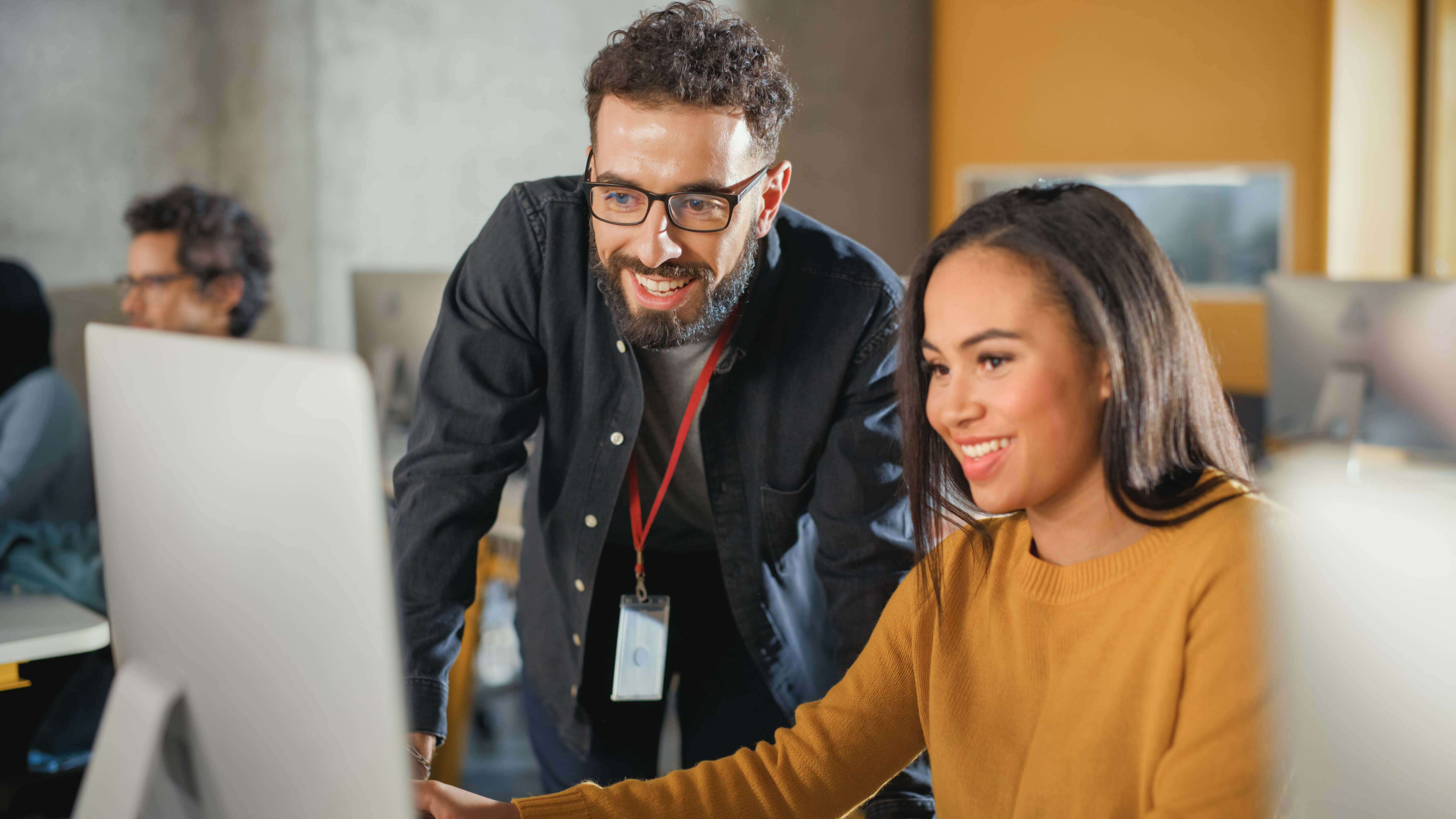 Smiling man in glasses leaning over to help a woman at a computer in an office setting.