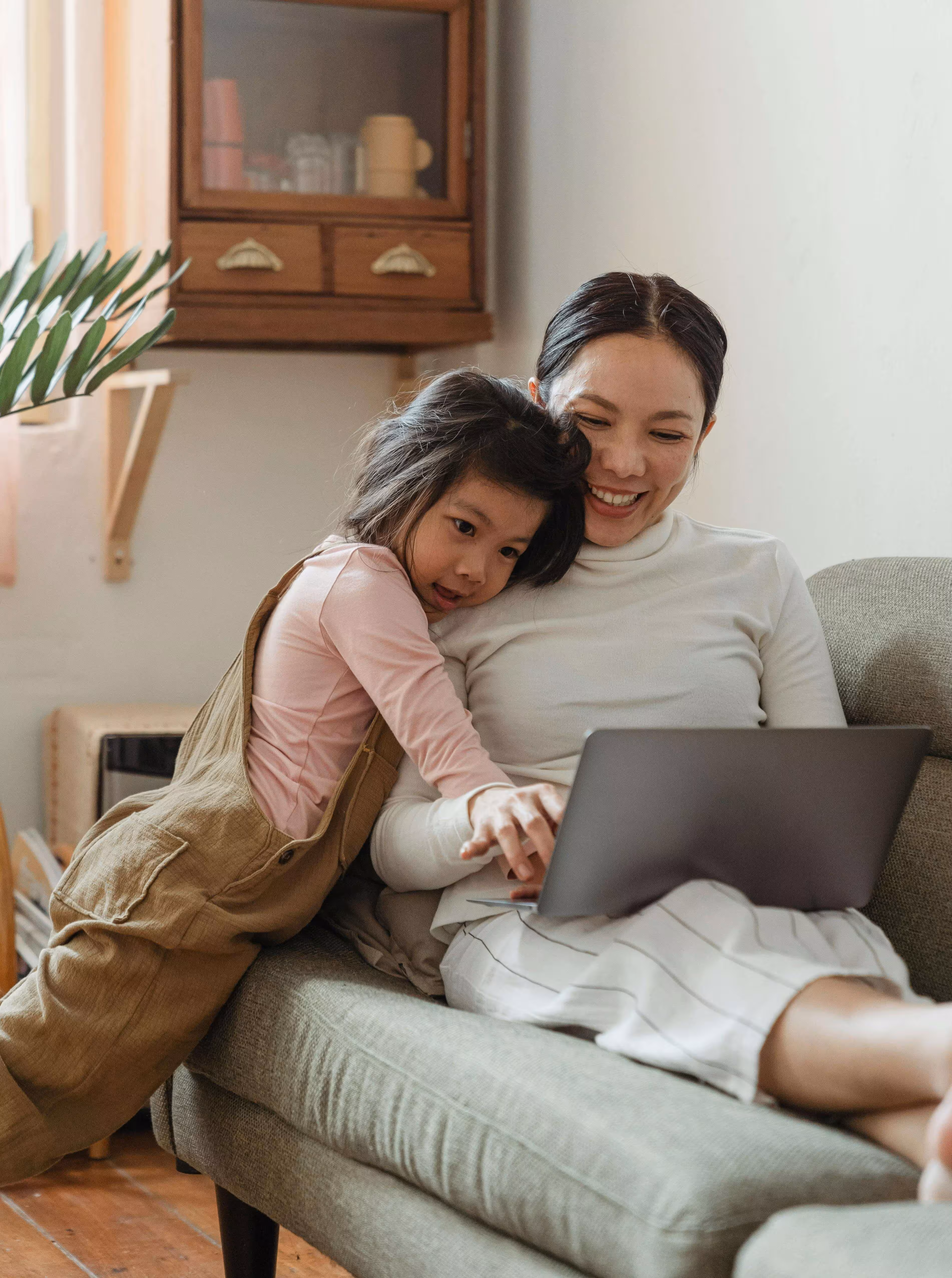 Smiling woman sitting on a couch using a laptop while a young girl leans on her shoulder and points at the screen.