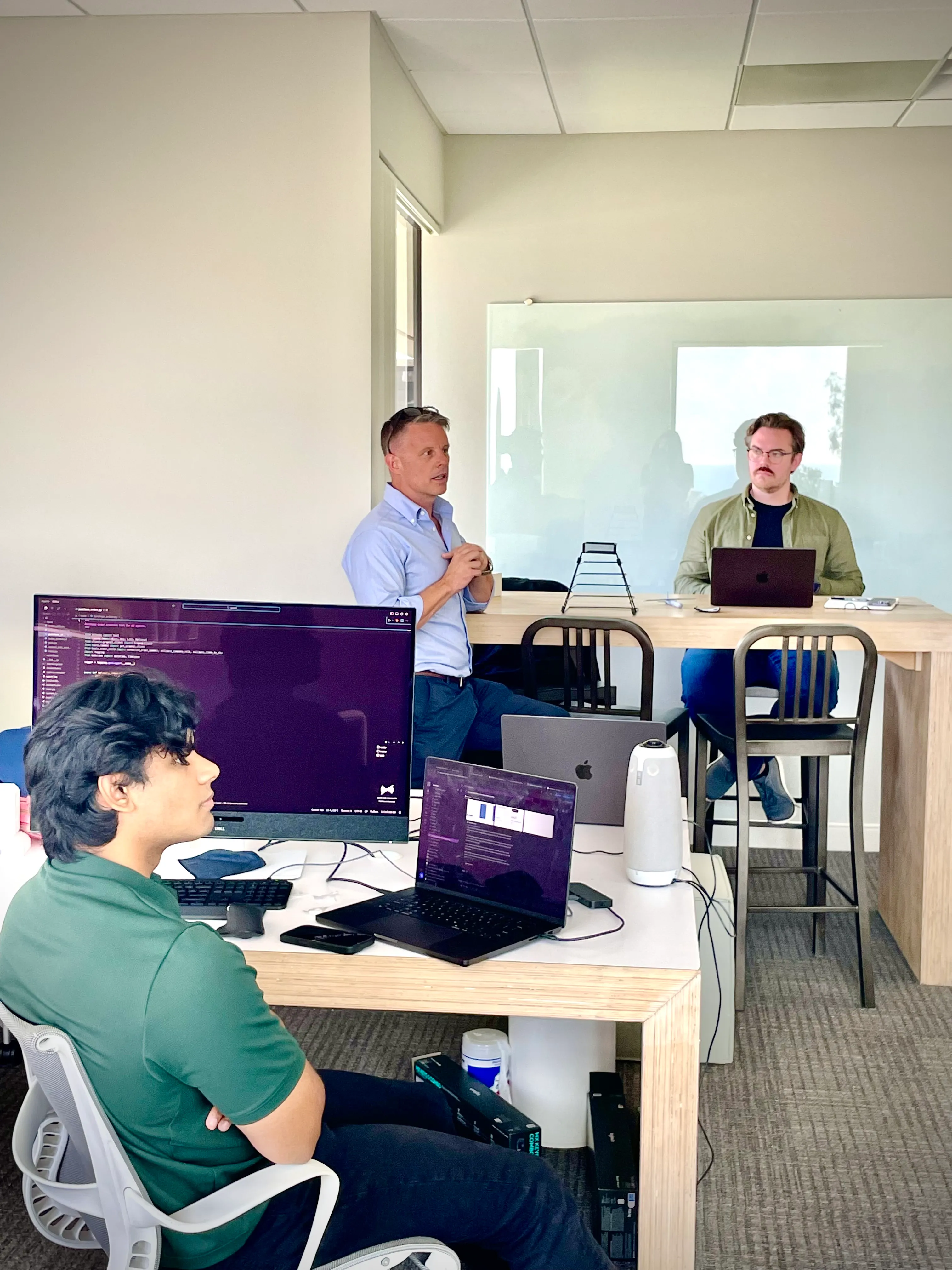 Three men in office discussing near laptops and monitor with code on screens.
