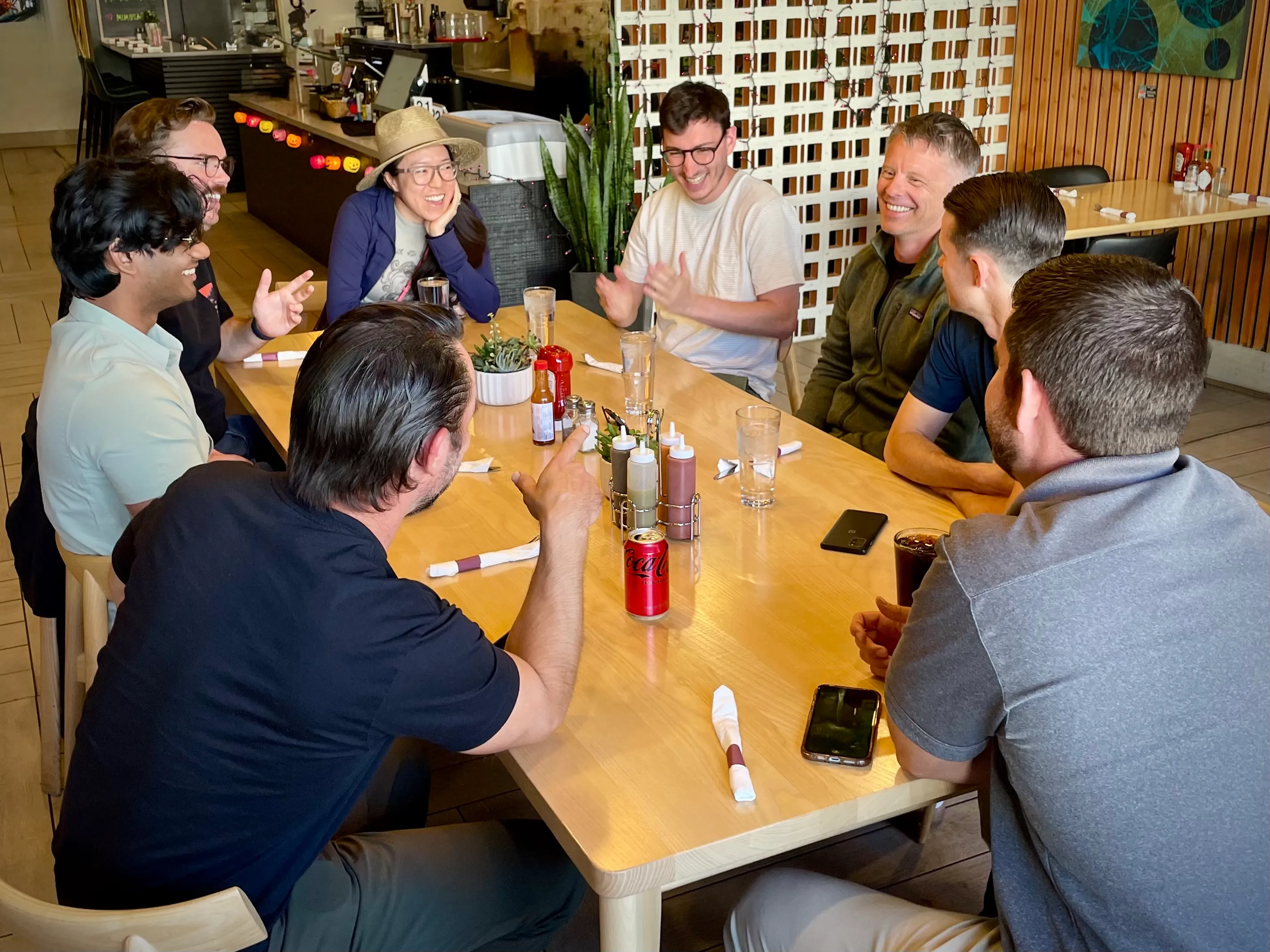 Eight adults sitting around a wooden table laughing and talking inside a casual restaurant.
