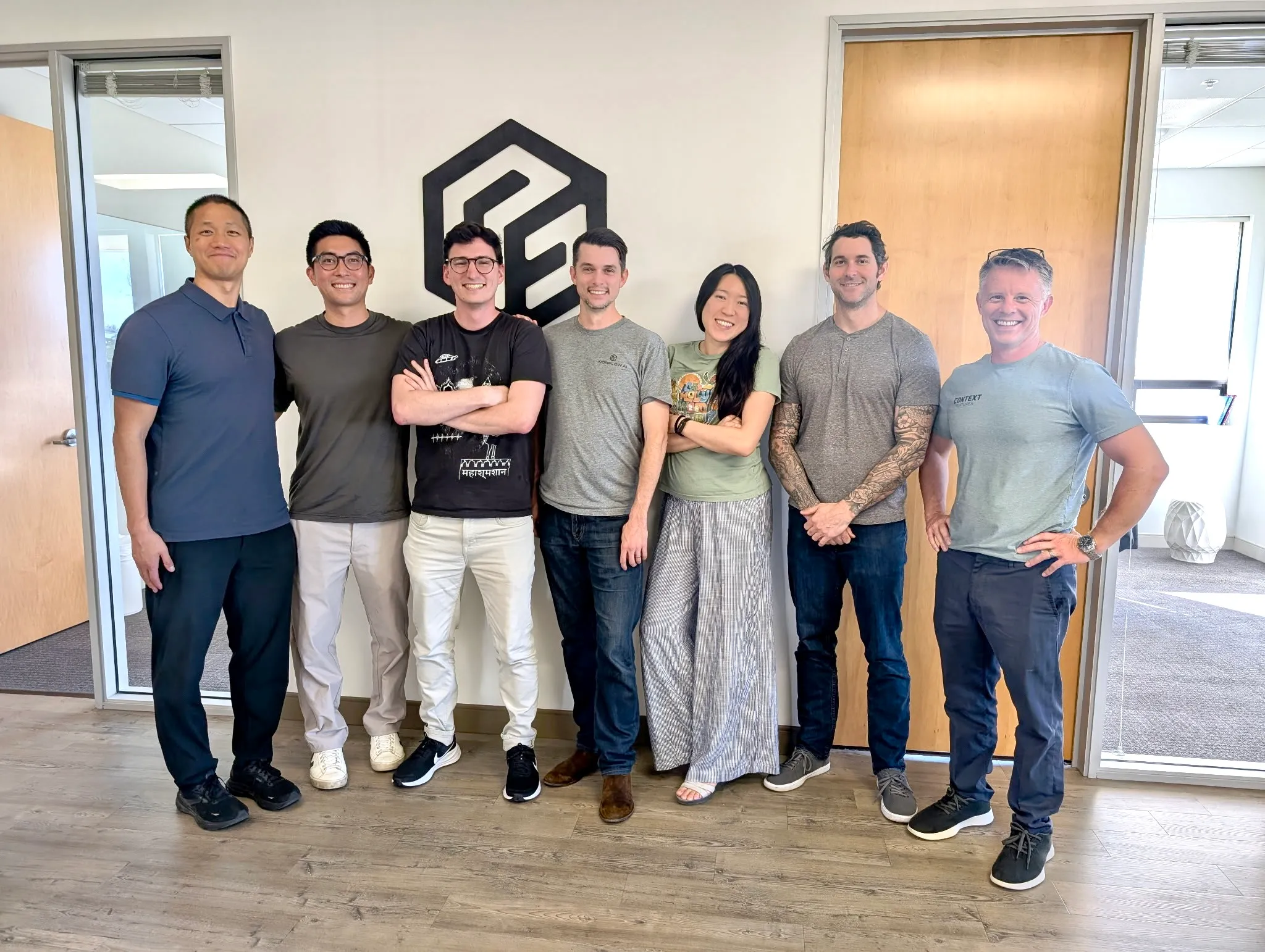 Seven people standing in an office in front of a wall with a hexagonal logo, smiling and posing.