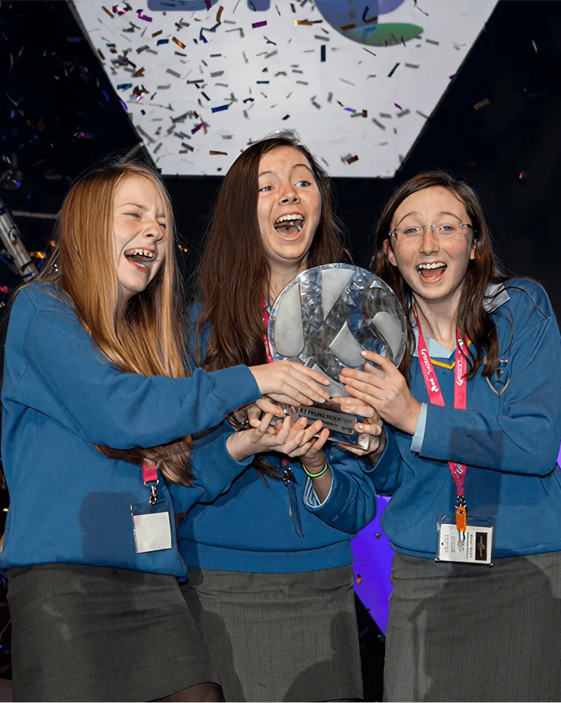 Three girls in blue uniforms accepting an award on stage with confetti falling.