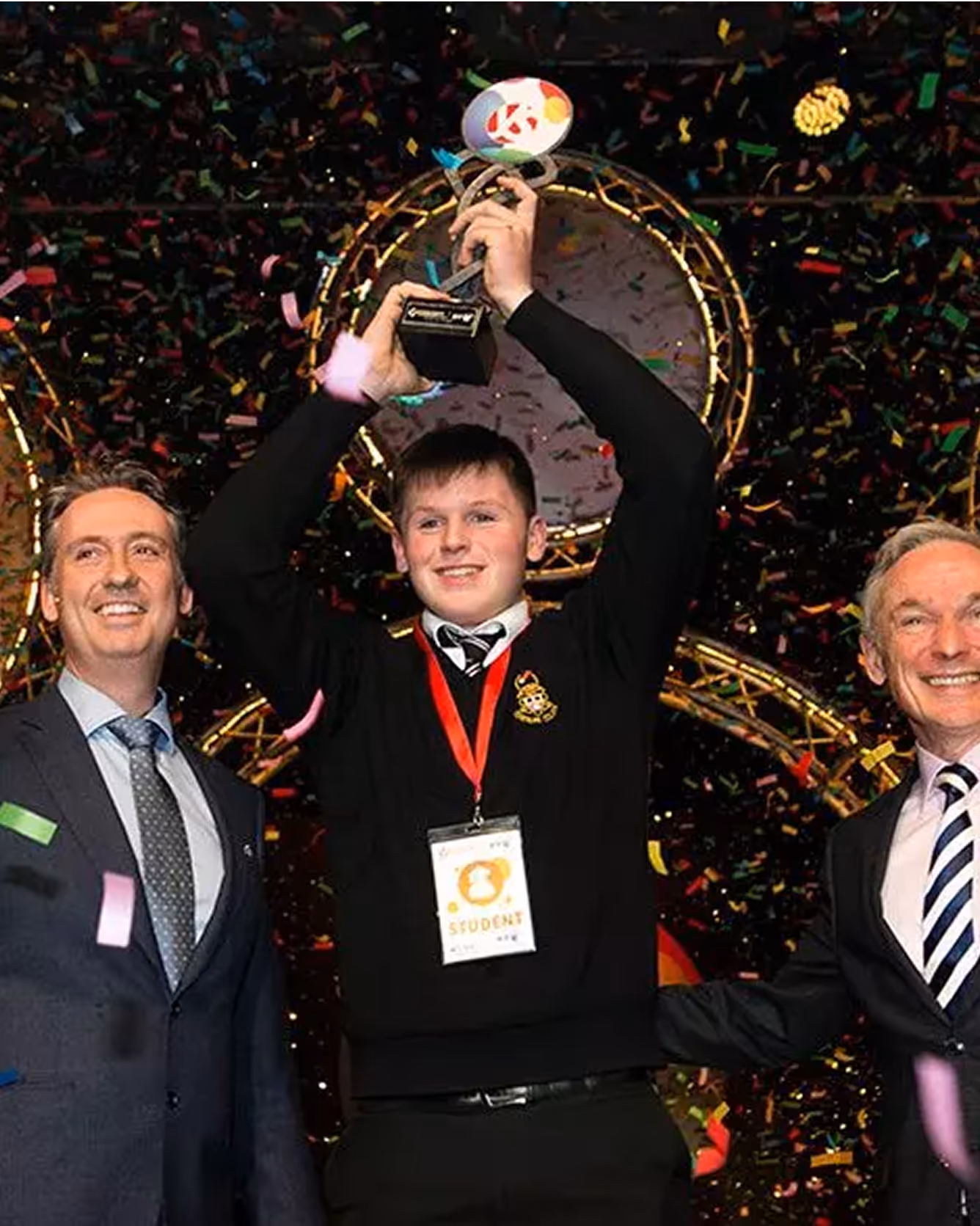 Student holding award trophy on stage with confetti, standing beside two men.