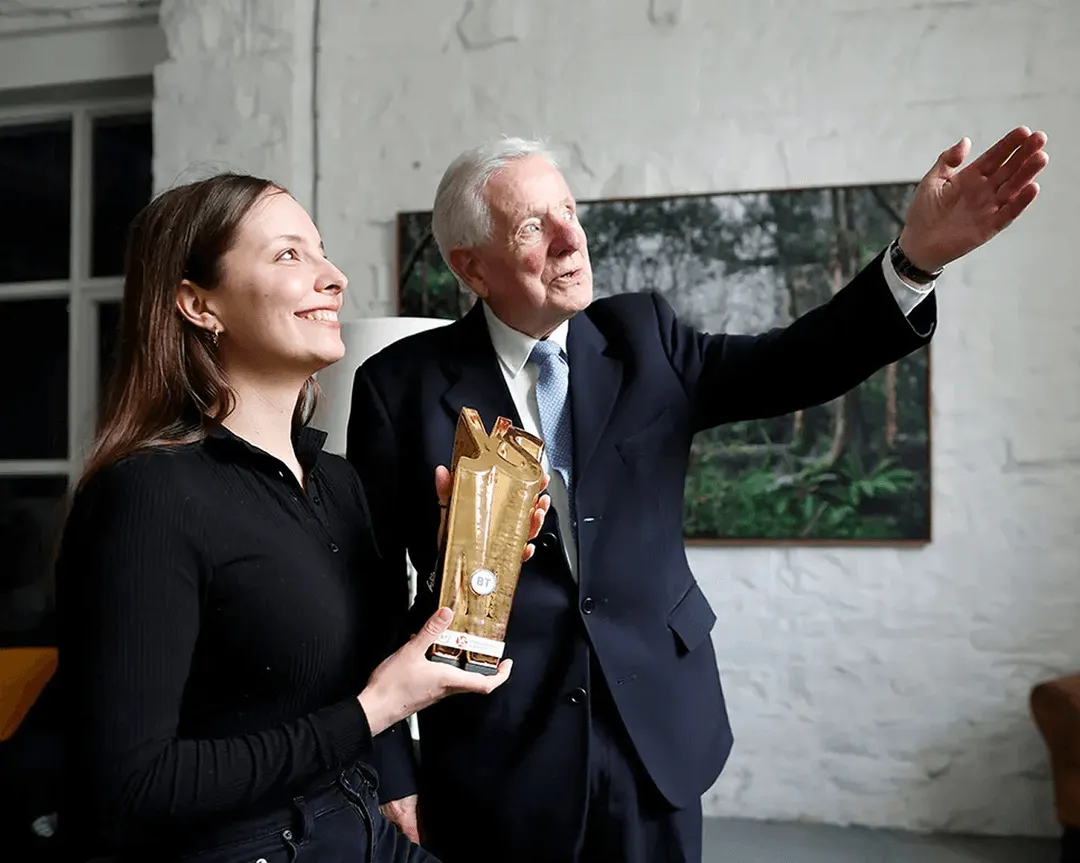 Smiling woman holding a gold award beside an older man in a suit gesturing ahead.
