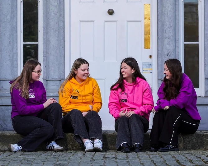 Four students wearing colorful hoodies sit on steps outside a building, chatting and smiling together.