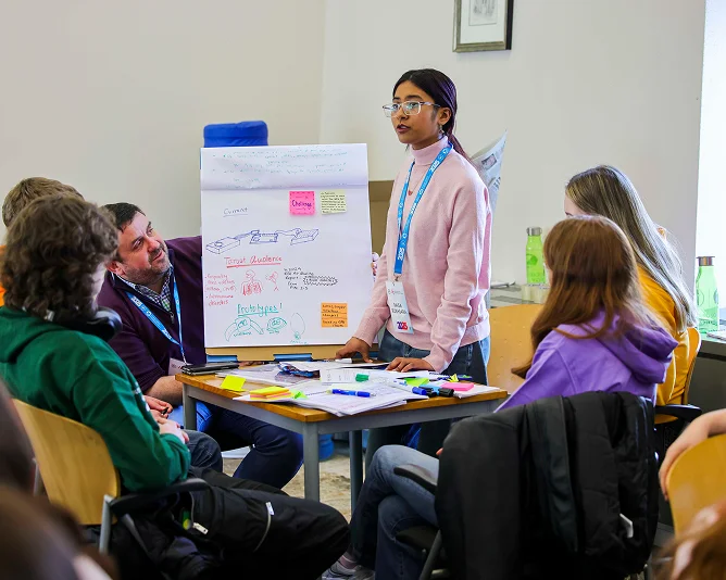 A student stands presenting a poster with notes and diagrams to a small group seated around a table during a classroom workshop.
