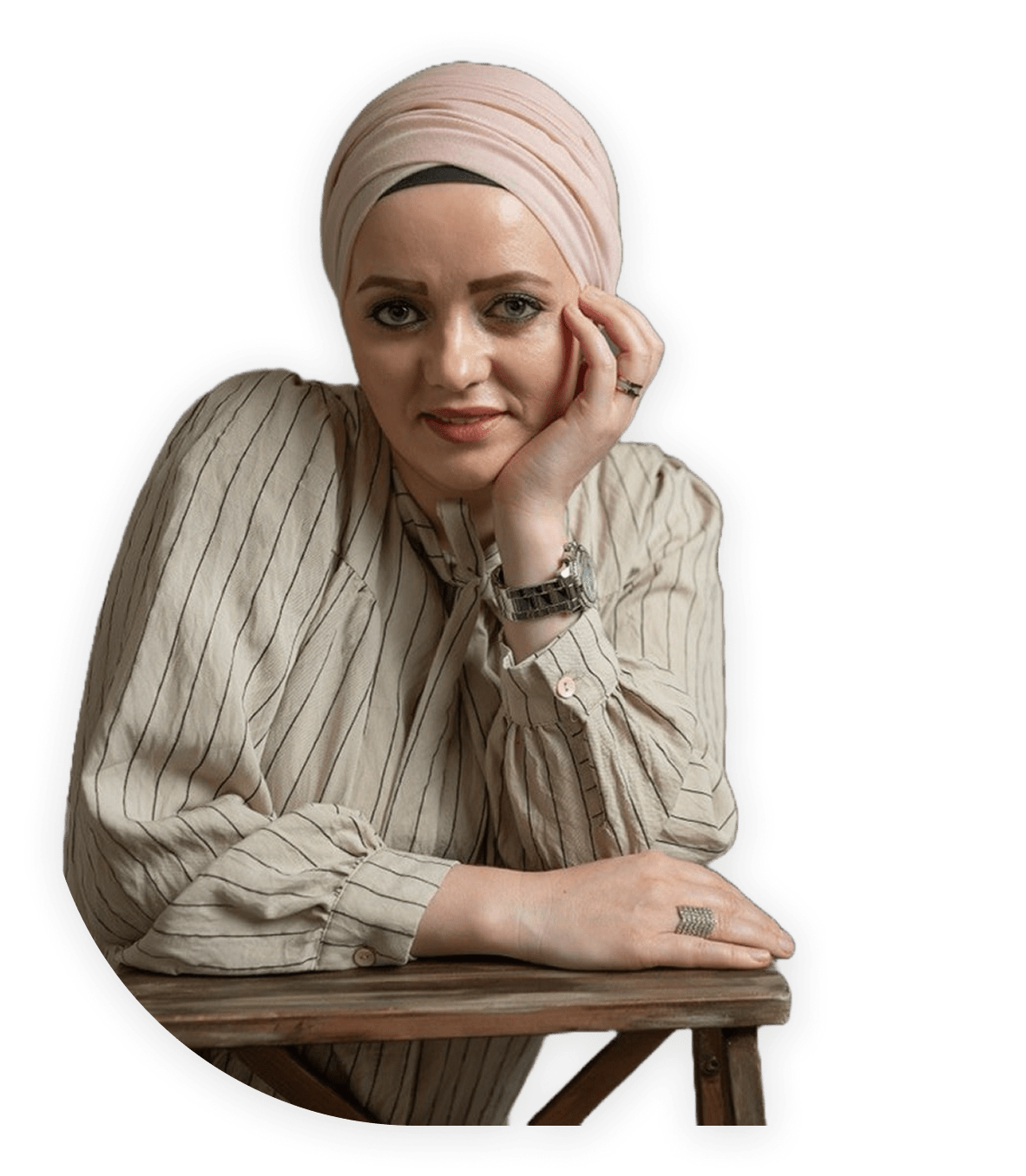 Woman wearing a light pink headscarf and striped beige blouse leaning on a wooden table, resting her face on one hand and looking at the camera.