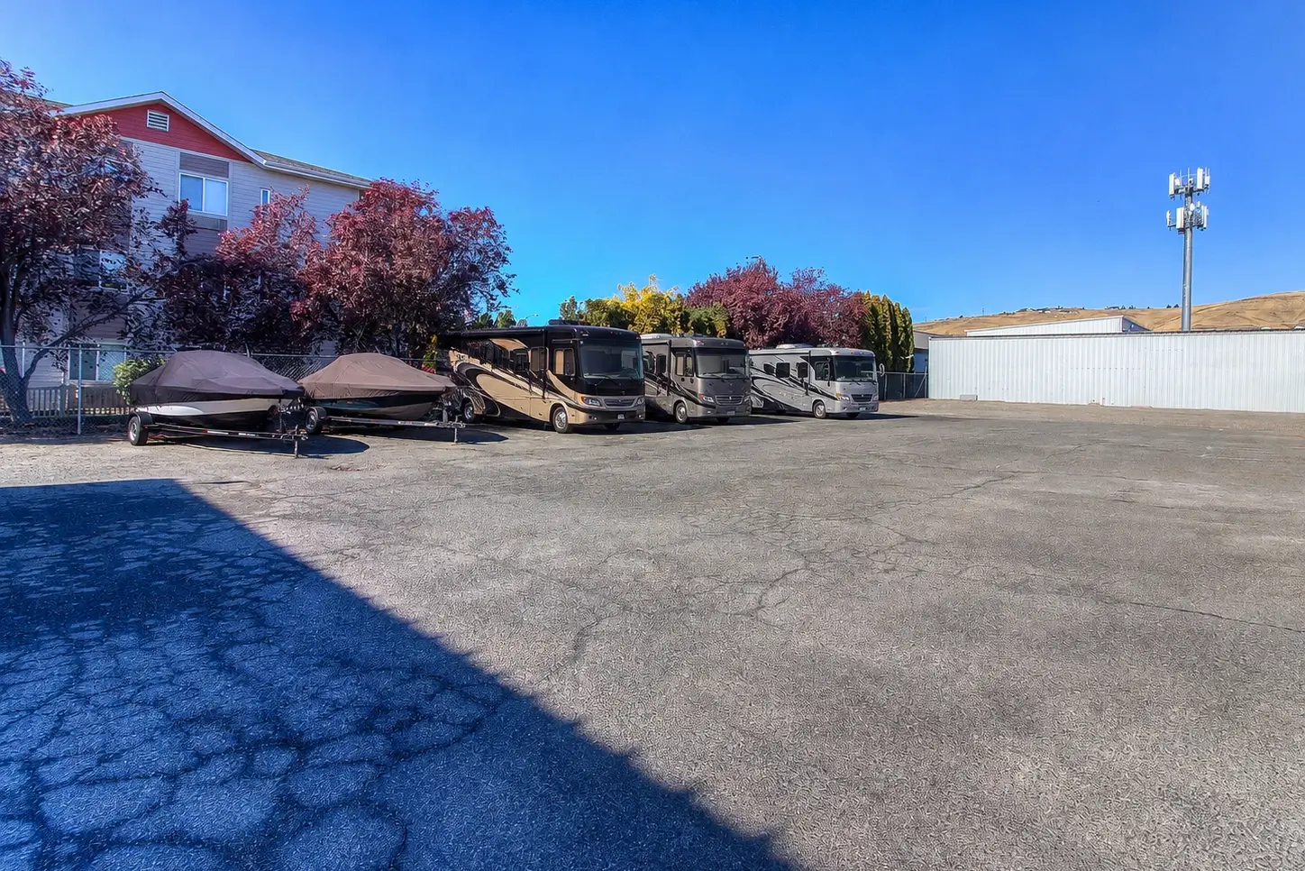 Outdoor parking lot with two covered boats on trailers and three large RVs parked side by side, surrounded by a fence with trees and buildings in the background under a clear blue sky.