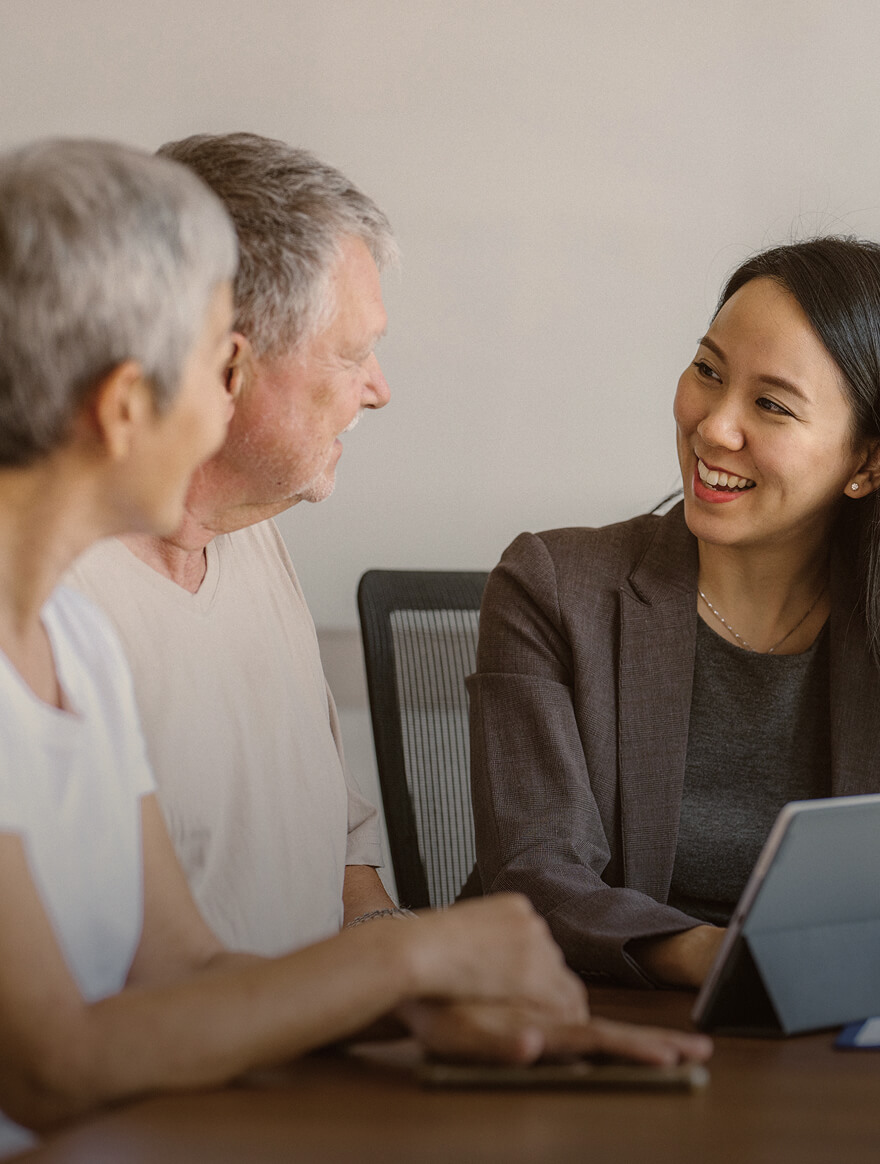 A smiling young woman in a blazer talks with two older adults seated at a table with a tablet device.