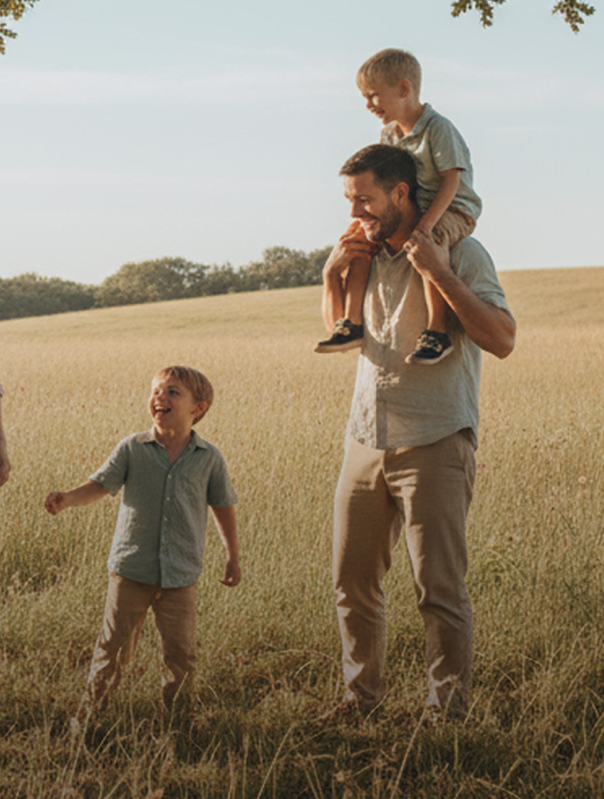 Man carrying a smiling child on his shoulders while another child stands nearby in a sunlit field.