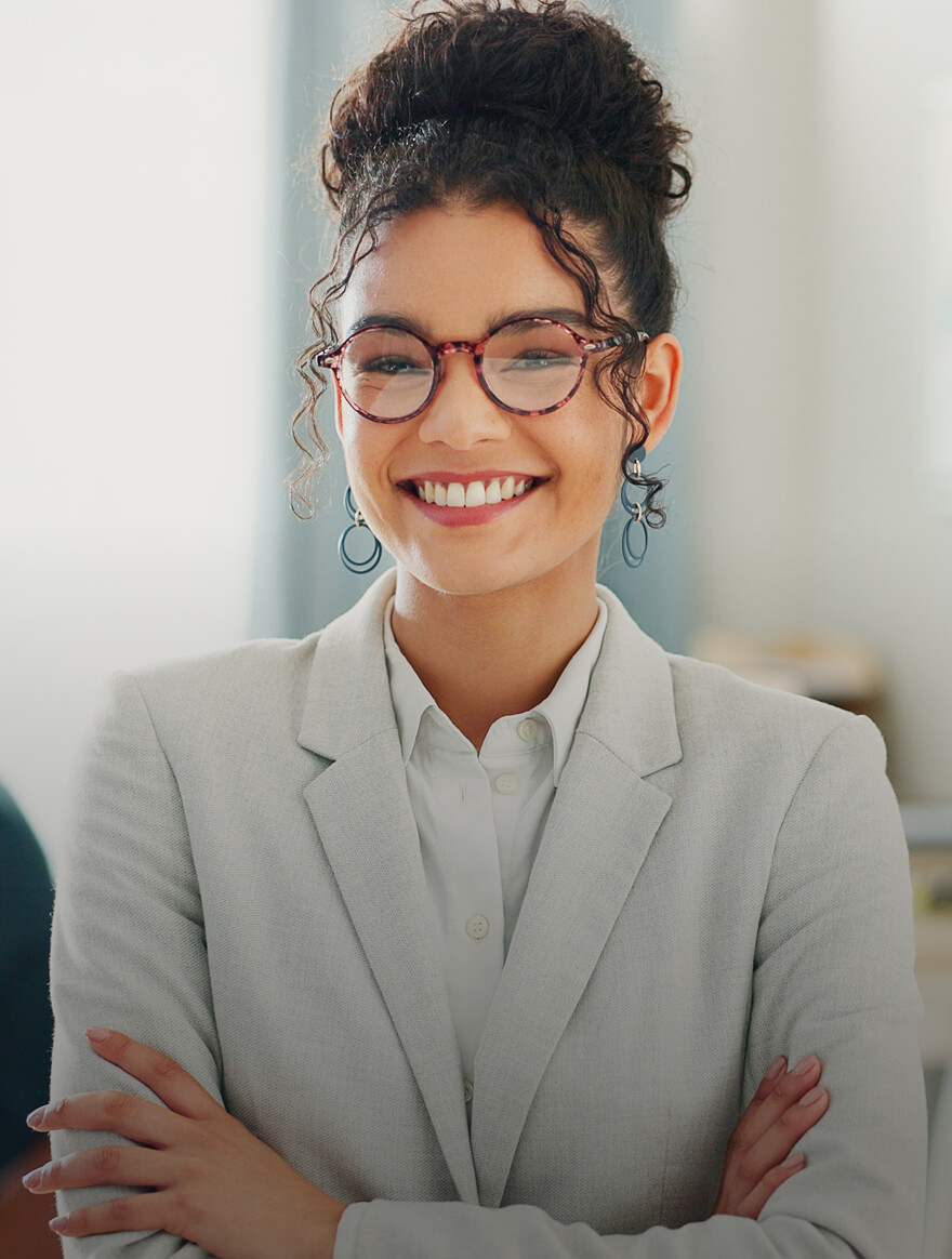 Smiling professional woman with curly hair in a bun, wearing glasses and a light gray blazer, standing with arms crossed.