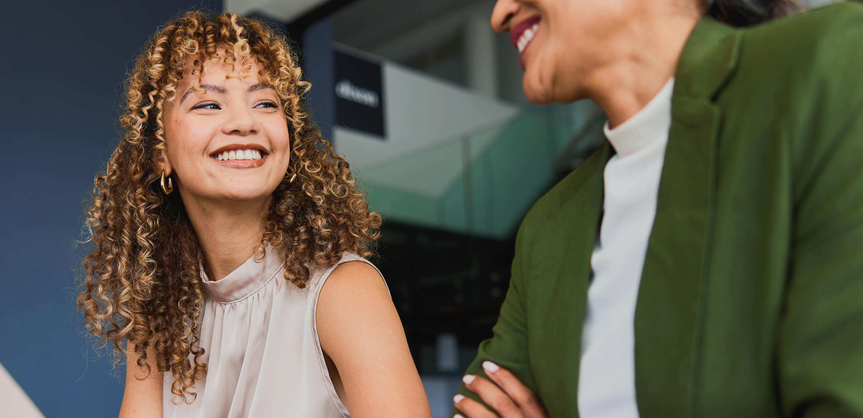 Two smiling women engaged in conversation, one with curly hair wearing a sleeveless blouse, the other in a green blazer.