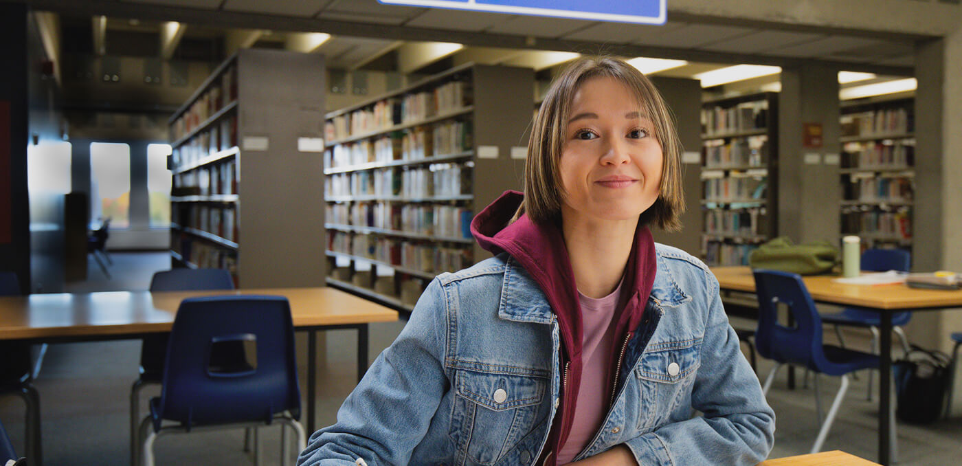 Young woman smiling while sitting at a table in a library with bookshelves and empty chairs in the background.