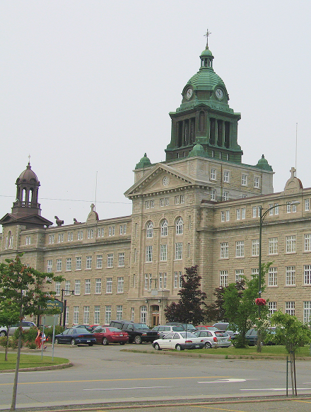 Large historic stone building with green clock tower and multiple windows, parked cars in front, and trees surrounding it.