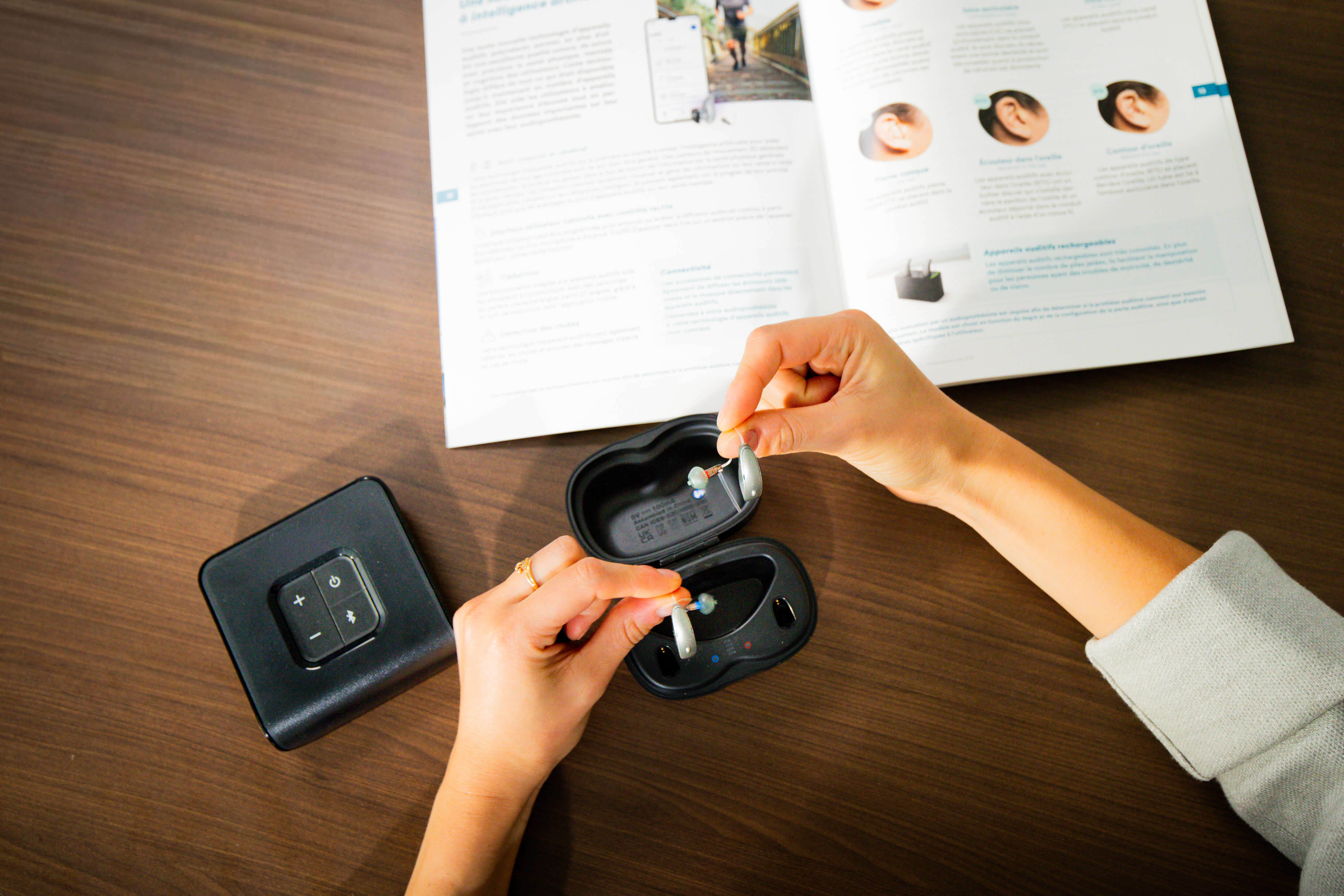 Person holding two hearing aids above a black charging case on a wooden table with an open magazine.