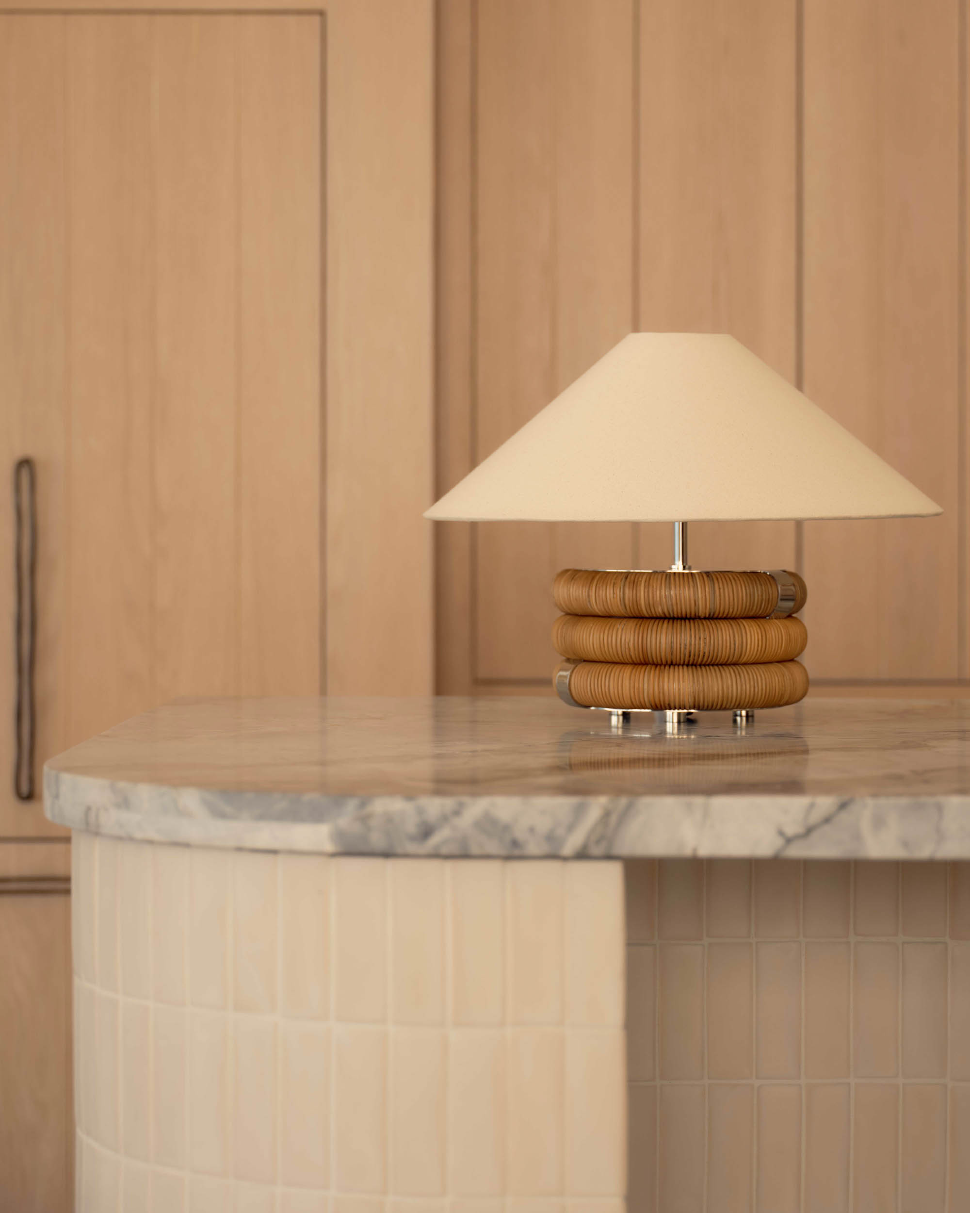 Table lamp with a beige conical shade and coiled wooden base on a marble countertop with light wood paneled cabinets in background.