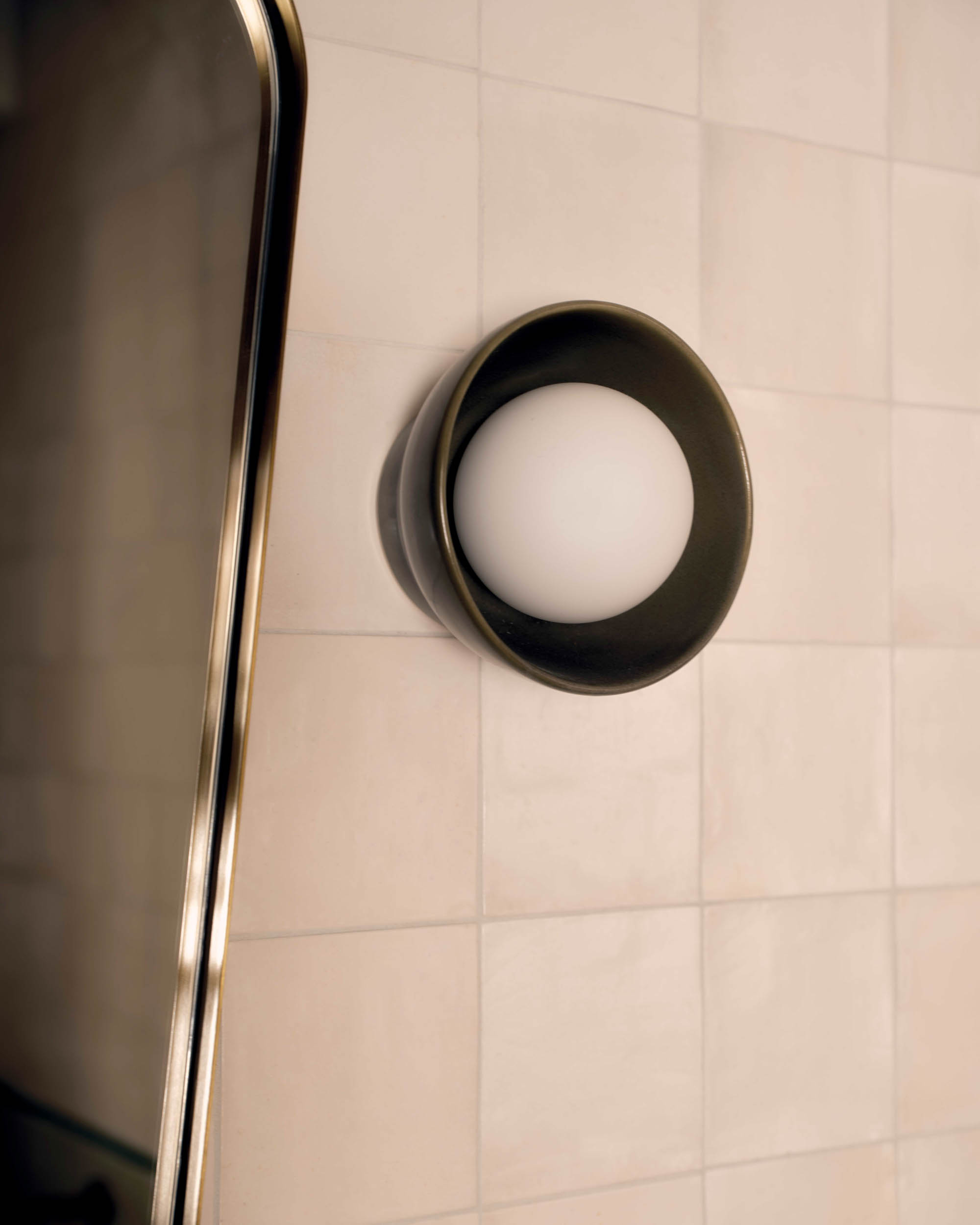Close-up of a round wall-mounted light fixture with a white globe on beige tiled wall next to mirrored edge.