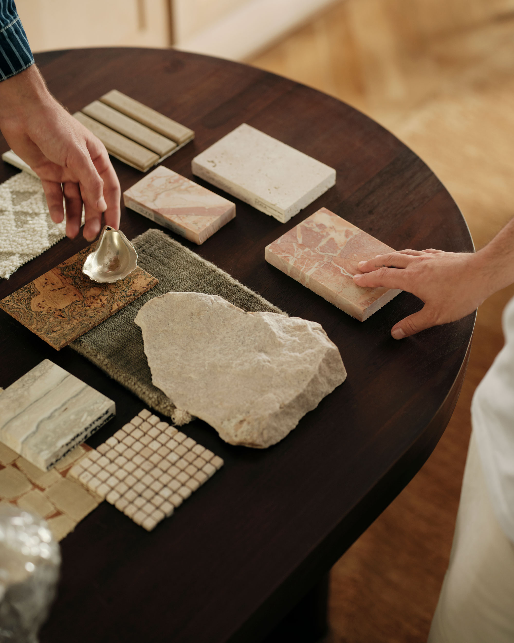 Two interior designers arranging various stone, tile, and fabric samples on a dark wooden table.