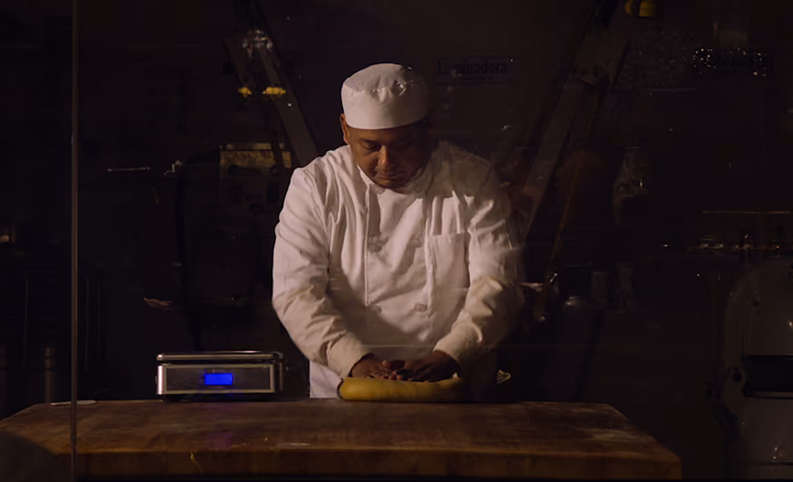 Chef in white uniform and hat kneading dough on a wooden table in a dimly lit kitchen.