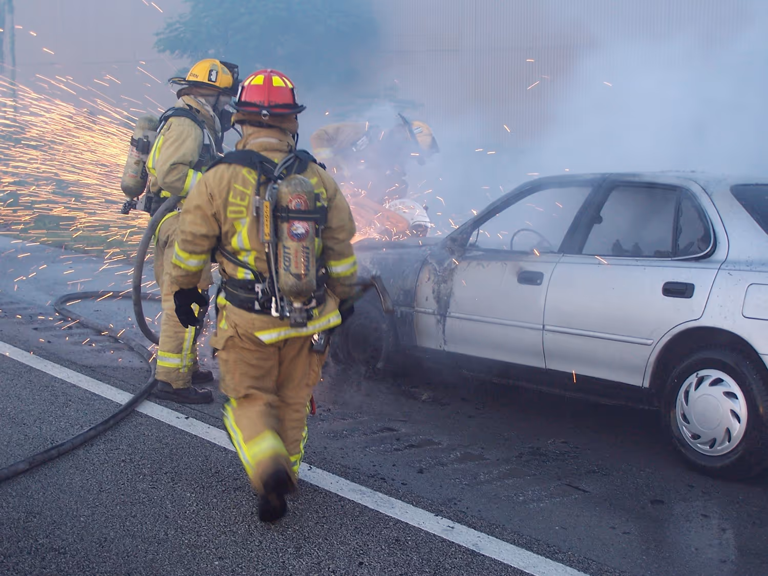 Delray Beach Fire Rescue firefighters extinguish a vehicle fire on the roadway as sparks and smoke rise from the car.