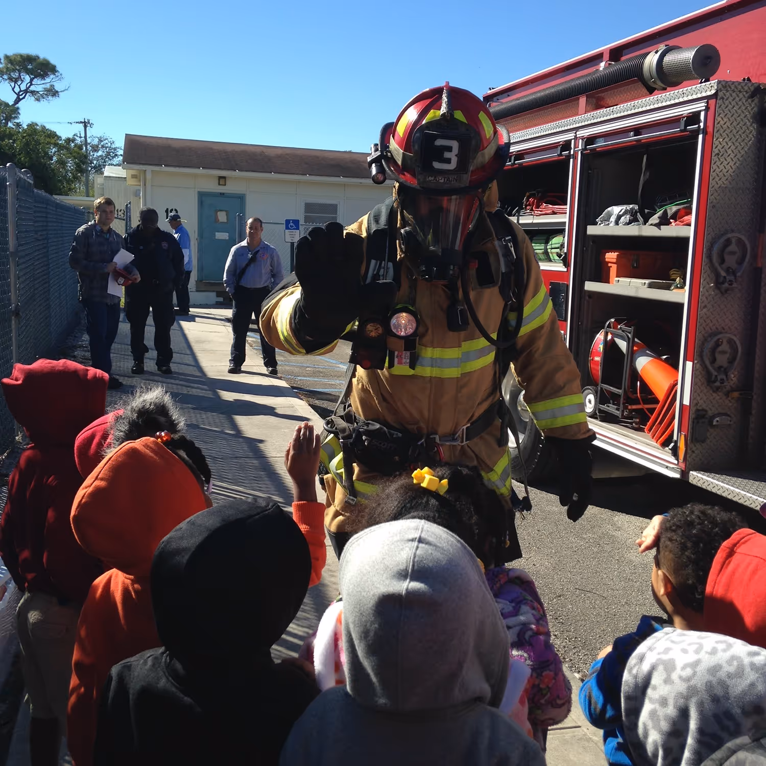Firefighter in full gear high-fiving young children during a school visit and safety demonstration.