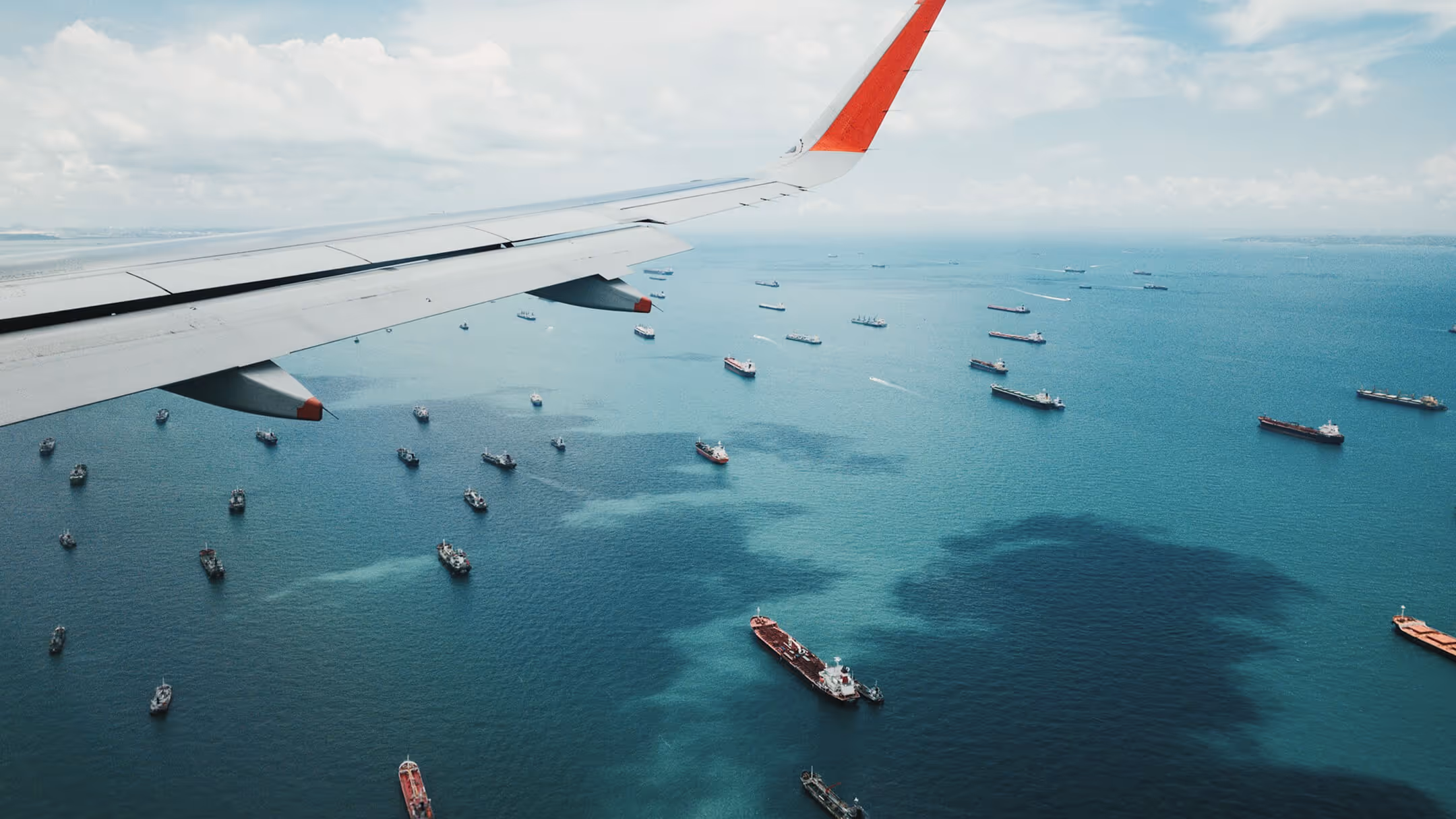 Airplane wing flying over a sea with numerous cargo ships and vessels scattered across the water.