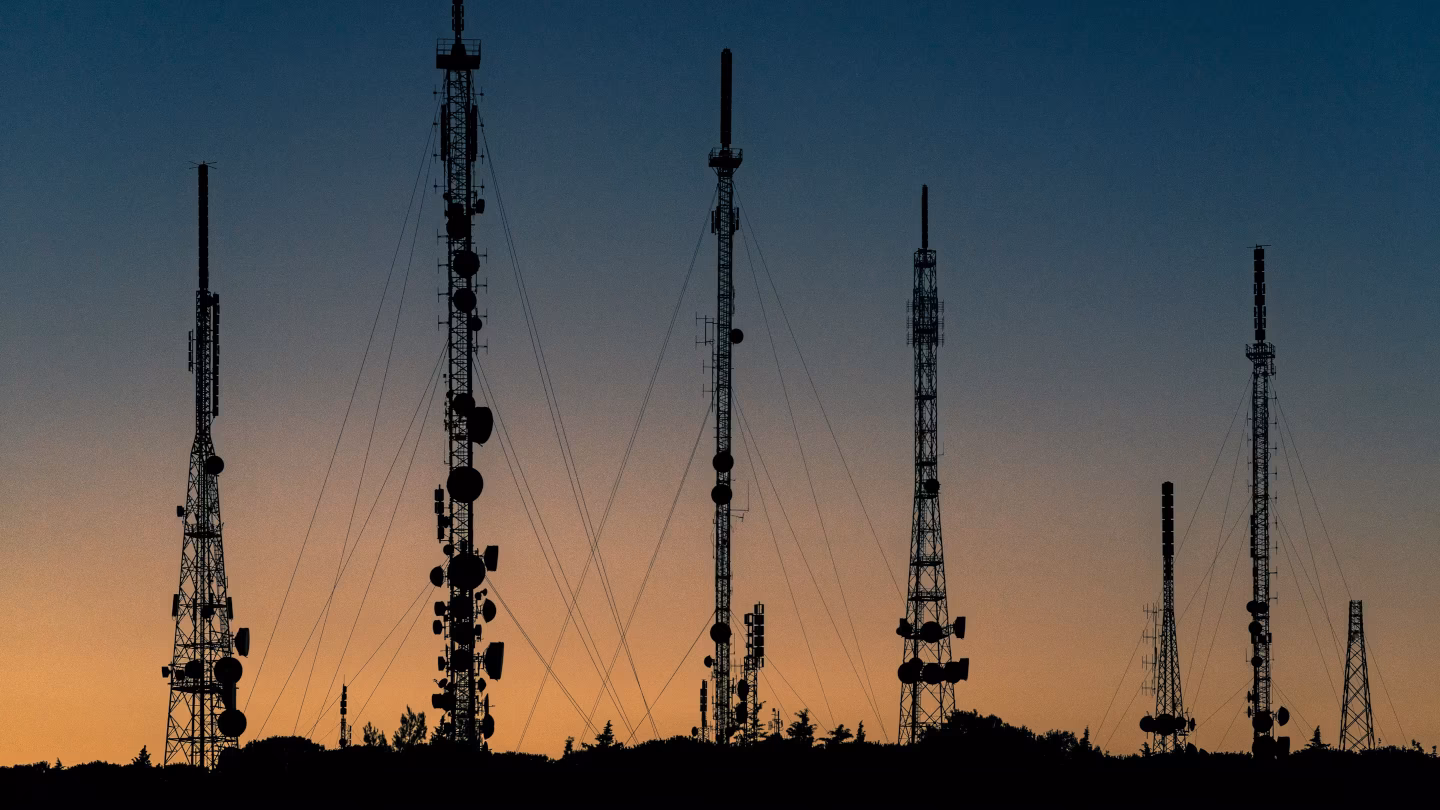 A skyline photograph of several communication towers set against a setting sun
