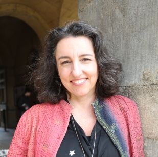 Smiling woman with dark curly hair wearing a coral jacket and black top standing against a stone wall.